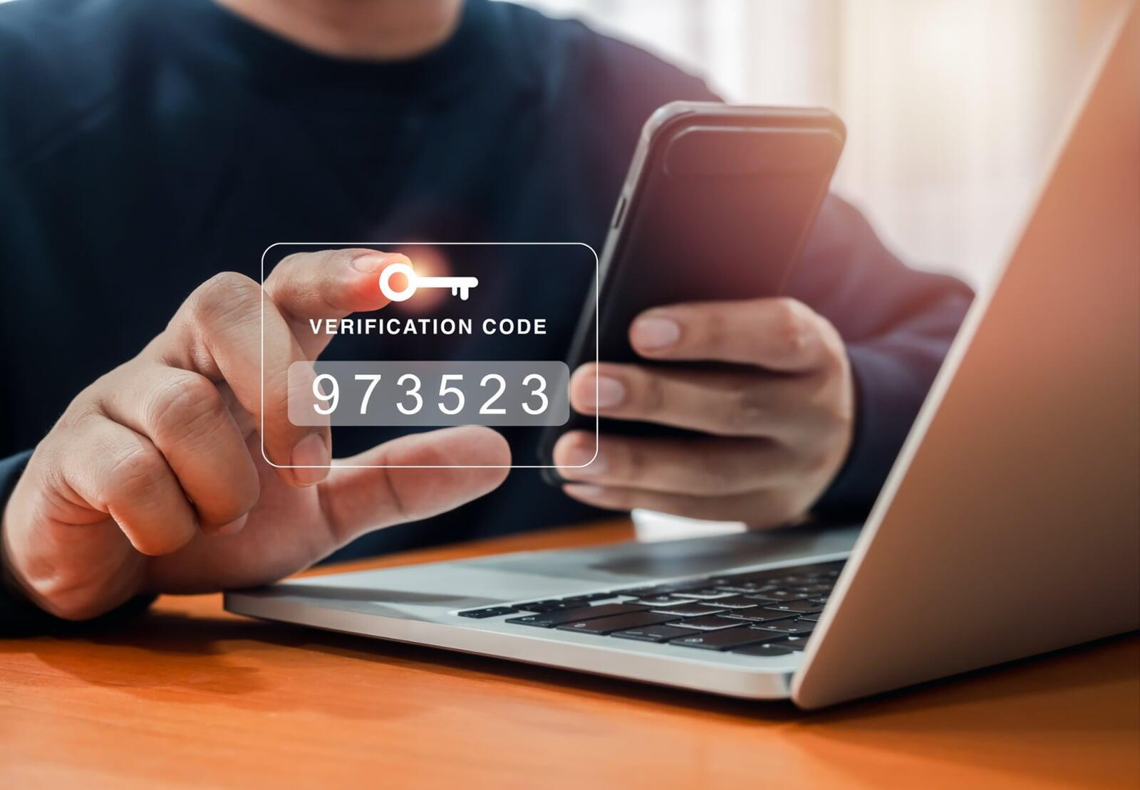 Hands using a laptop and phone displaying a "VERIFICATION CODE" for Two-Factor Authentication Methods (2FA).