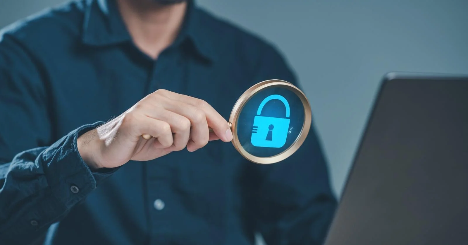 Man using a magnifying glass to view a glowing blue lock icon on a laptop, symbolizing a deep scan to Detect Hidden Malware.