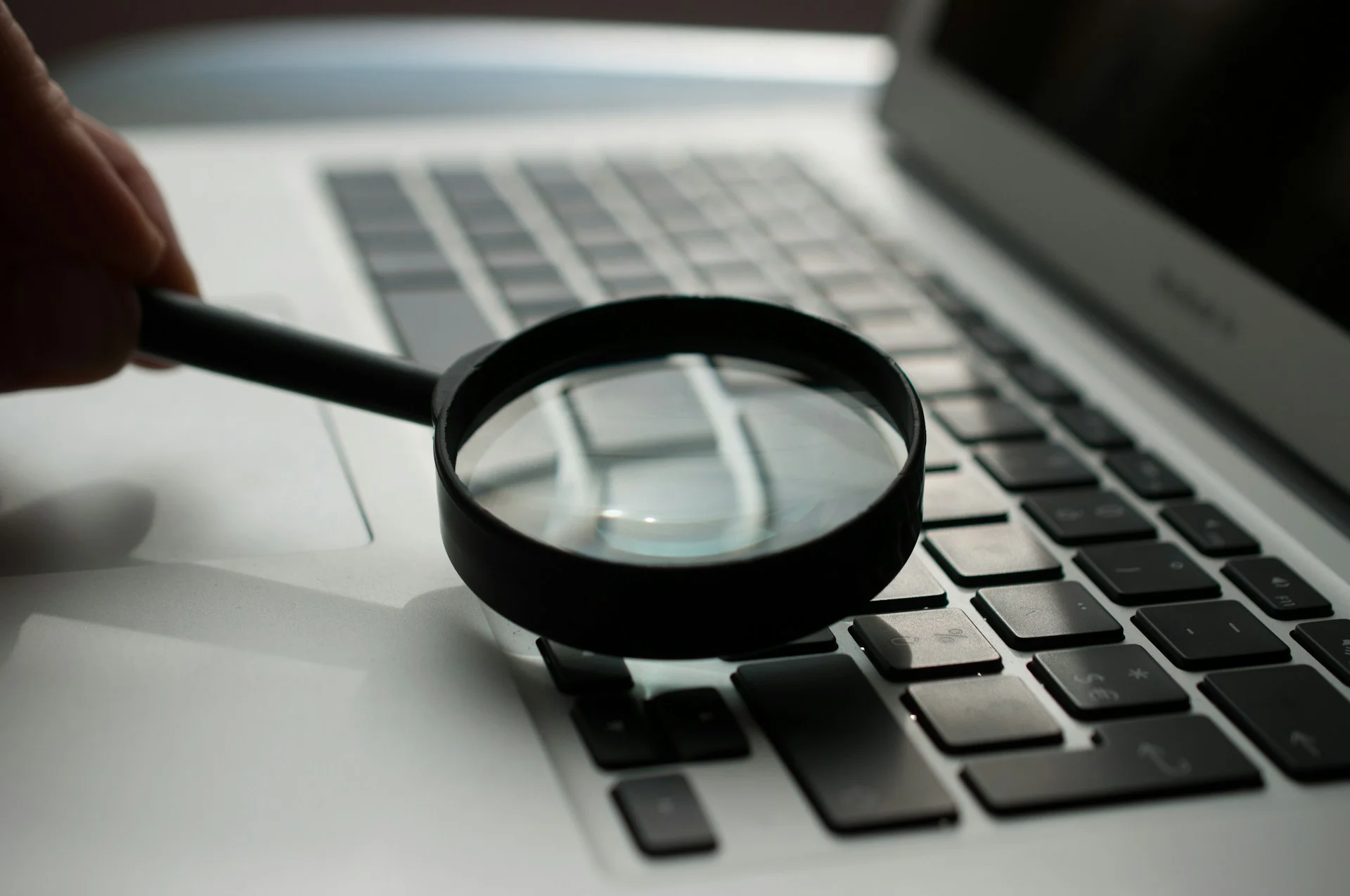 A hand holding a magnifying glass over a laptop keyboard, representing detailed technical audits and routine maintenance for a Small Business IT Checklist.